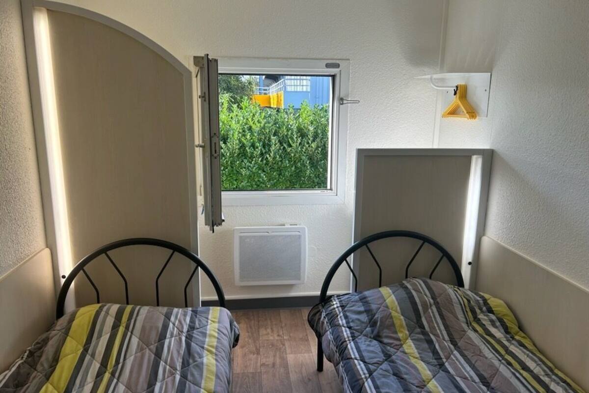 Una habitación pequeña con dos camas, una ventana que da a zonas verdes, un radiador y una lámpara fijada a la pared.
