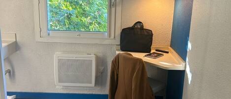 A hotel room with a bed, a window with a view of trees, a television mounted on the wall, and a small table with a bag and a remote control.
