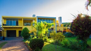 A yellow building with a paved driveway and a well-maintained garden with various plants and a small tree. - Room in Villa - Comfortable and Welcoming Suite With Terrace Overlooking the Lake (Antananarivo)