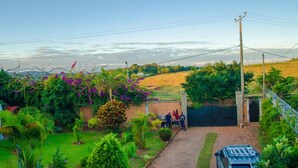 A rural property with a paved driveway leading to a gated entrance, surrounded by lush greenery and flowering plants. - Room in Villa - Comfortable and Welcoming Suite With Terrace Overlooking the Lake (Antananarivo)