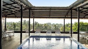 An indoor swimming pool with a view of palm trees, surrounded by white chairs and tables under a metal roof. -  La Baga Beach Hotel (Calangute)