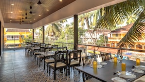 Een restaurant met terras, houten tafels en stoelen, met uitzicht op palmbomen en gebouwen.