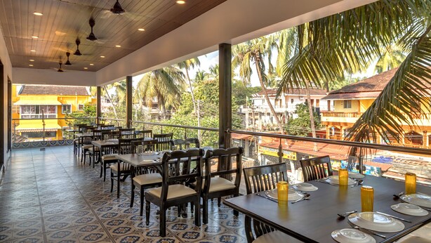 A restaurant with outdoor seating, wooden tables, and chairs, overlooking palm trees and buildings. -  La Baga Beach Hotel (Calangute)