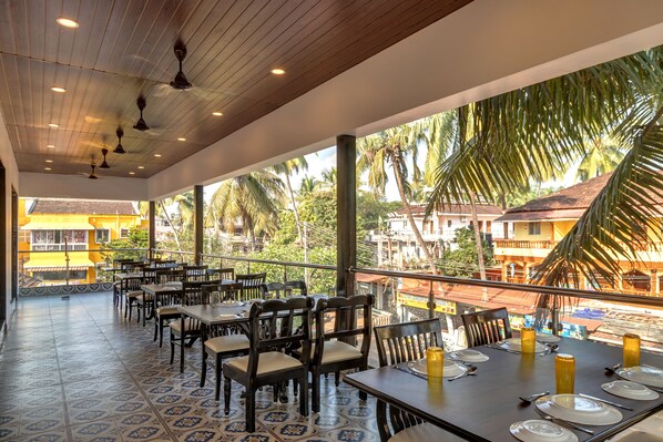 Un restaurant doté d’une terrasse, de tables et de chaises en bois, offrant une vue sur les palmiers et les bâtiments environnants.