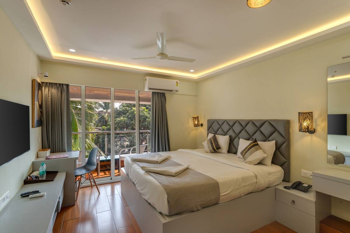 Een moderne hotelkamer met een groot bed, een bureau met stoel, een televisie en een balkon met uitzicht op palmbomen.