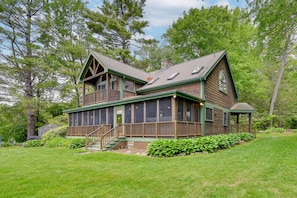 A two-story wooden cabin with a covered porch, large windows, and a stone chimney.