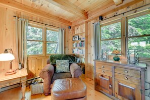 A cozy cabin interior with a wooden interior, a sofa with cushions, a small table with a lamp, a bookshelf, and large windows offering a view of trees.