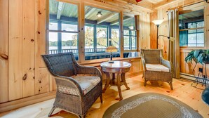 A wooden cabin interior with wicker furniture, large windows, and a view of the outdoors. - Lakefront Westhampton Cabin w/ Beach + Kayaks! (Westhampton)