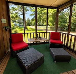 A covered outdoor seating area with wicker furniture and red cushions, a small table with potted plants, and a view of a green landscape.