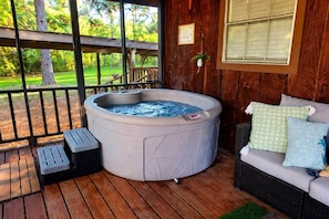 A hot tub on a wooden deck with a view of a garden, a wicker sofa with cushions, and a small trash bin.