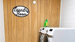 A laundry room with a wooden wall, a sign reading 'Laundry Room,' and two stacked front-loading washing machines. - The Gator Den at Caddo Lake / Big Lake - Hot Tub - Boat Ramp - Outdoor Games (Karnack)