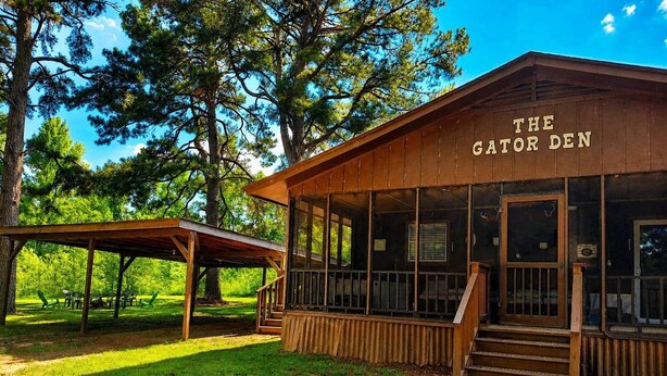 A wooden building labeled 'The Gator Den' with a covered outdoor seating area and a staircase leading to the entrance. - The Gator Den at Caddo Lake / Big Lake - Hot Tub - Boat Ramp - Outdoor Games (Karnack)