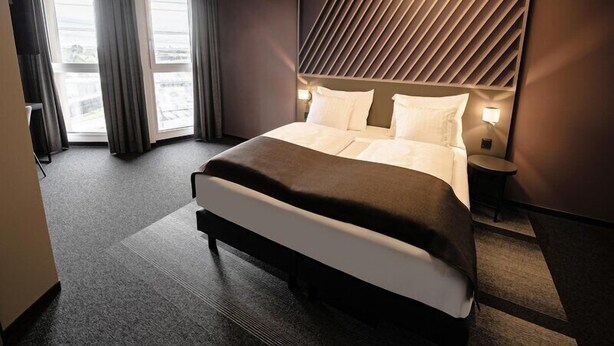 Una habitación de hotel moderna con una cama grande, una mesita de noche, una lámpara de pared y vistas a la ciudad desde la ventana.