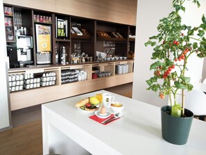 Una cafetería con mostrador blanco, una máquina de café y una maceta con tomates.