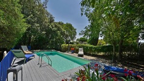 A rectangular swimming pool with a wooden deck, surrounded by lush greenery and trees.