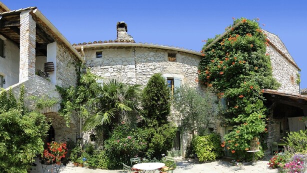 A stone building with a round tower, surrounded by lush greenery and flowering plants.