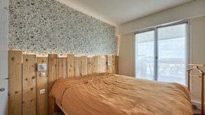 Une chambre à coucher avec une tête de lit en bois, un lit recouvert d’une couverture marron, une fenêtre donnant sur des immeubles et une armoire en bois.