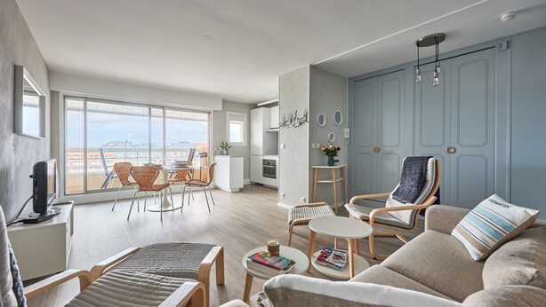 Un salon moderne avec un canapé, des fauteuils, une petite table et un téléviseur. Une grande fenêtre offre une vue sur la ville.