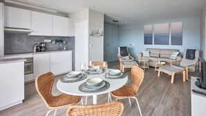 Une cuisine moderne avec des meubles blancs, un coin repas avec des chaises en osier et un salon avec un canapé et des fauteuils.