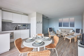 Eine moderne Küche mit weißen Schränken, eine Essecke mit Korbstühlen und ein Wohnzimmer mit einer Couch und Sesseln.