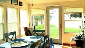 A dining area with a table set for four, surrounded by chairs, with a view of a green lawn through the door. - Beautiful Huron Bay sunsets from your own private beach. (Skanee)