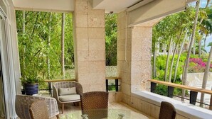 A sunlit outdoor seating area with wicker furniture and a glass table, surrounded by stone walls and large windows overlooking lush greenery. - Srvittinivillas Aptla F1 Best Loc and View / Lux / Altos D Chavon/ Cdc (La Romana)