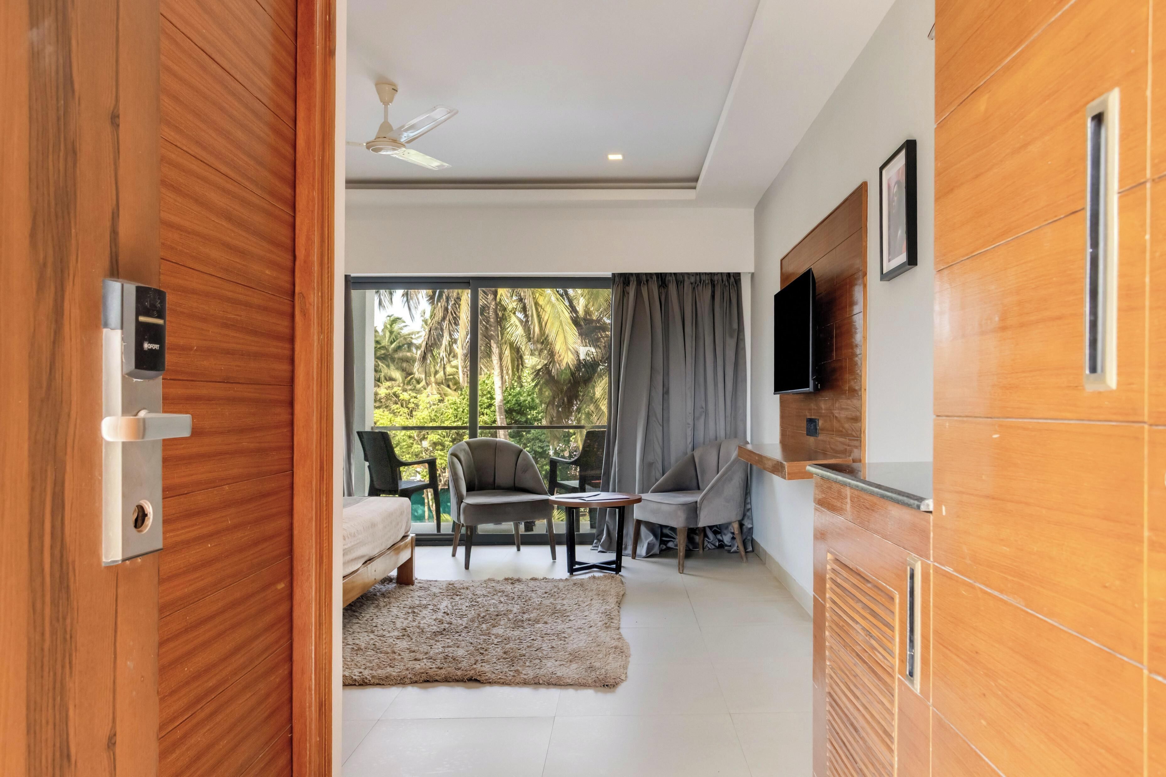 A modern room with a bed, two chairs, a small table, a TV, and a balcony with a view of trees.