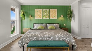 Un dormitorio con pared verde, cama con colcha floral gris y blanca, dos almohadas verdes y un banco.