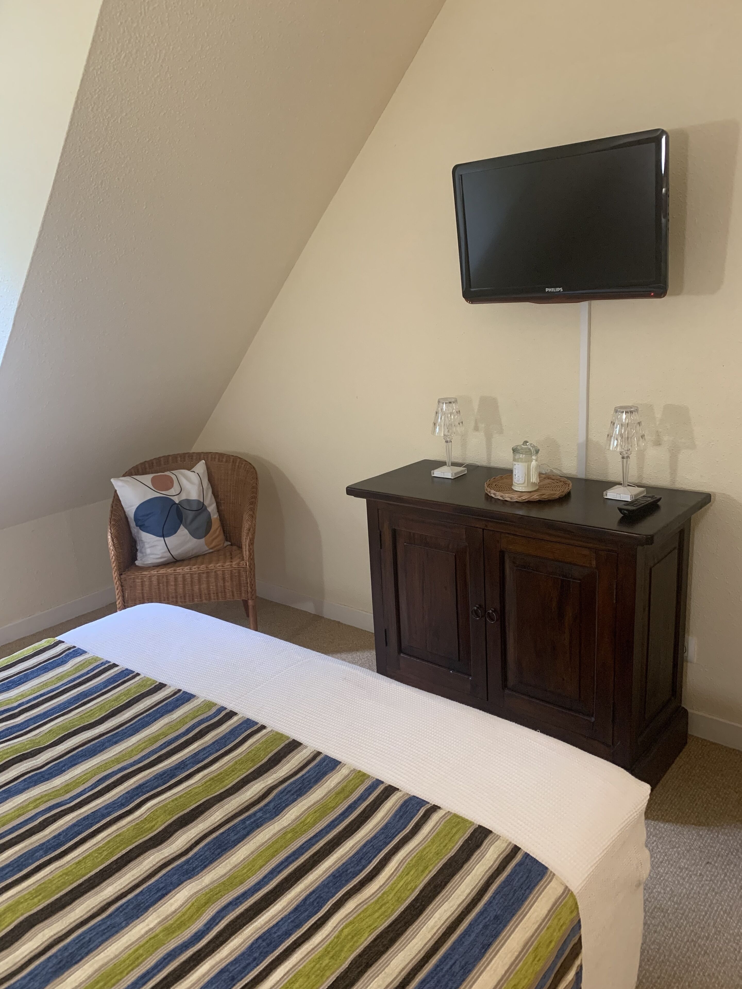 Une chambre d’hôtel avec un lit, une chaise en osier, une petite table avec deux lampes et un téléviseur fixé au mur.