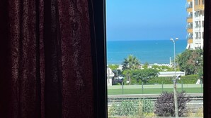 Vista a través de una ventana con cortina, mostrando un paisaje costero con cielo despejado, un edificio con balcones y una zona verde con árboles.
