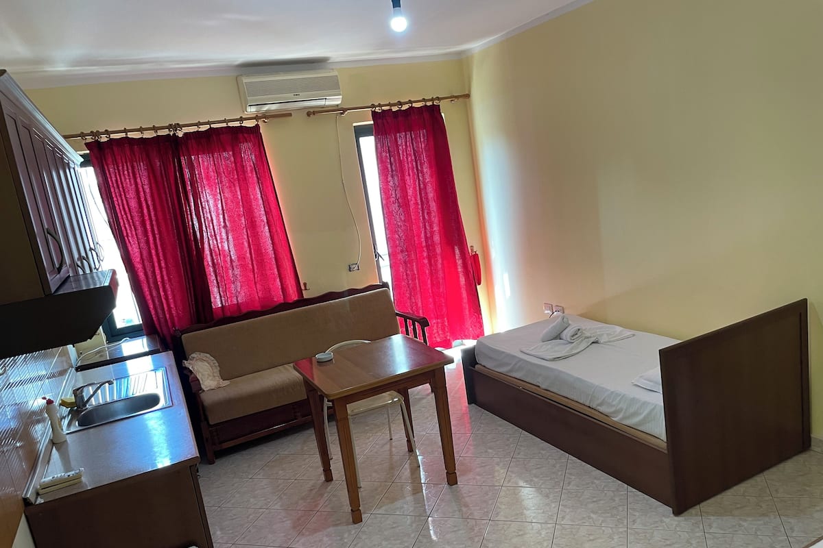 Quarto de hotel compacto com uma cama, um sofá, uma mesa pequena, uma escrivaninha com computador e uma janela com cortinas vermelhas.