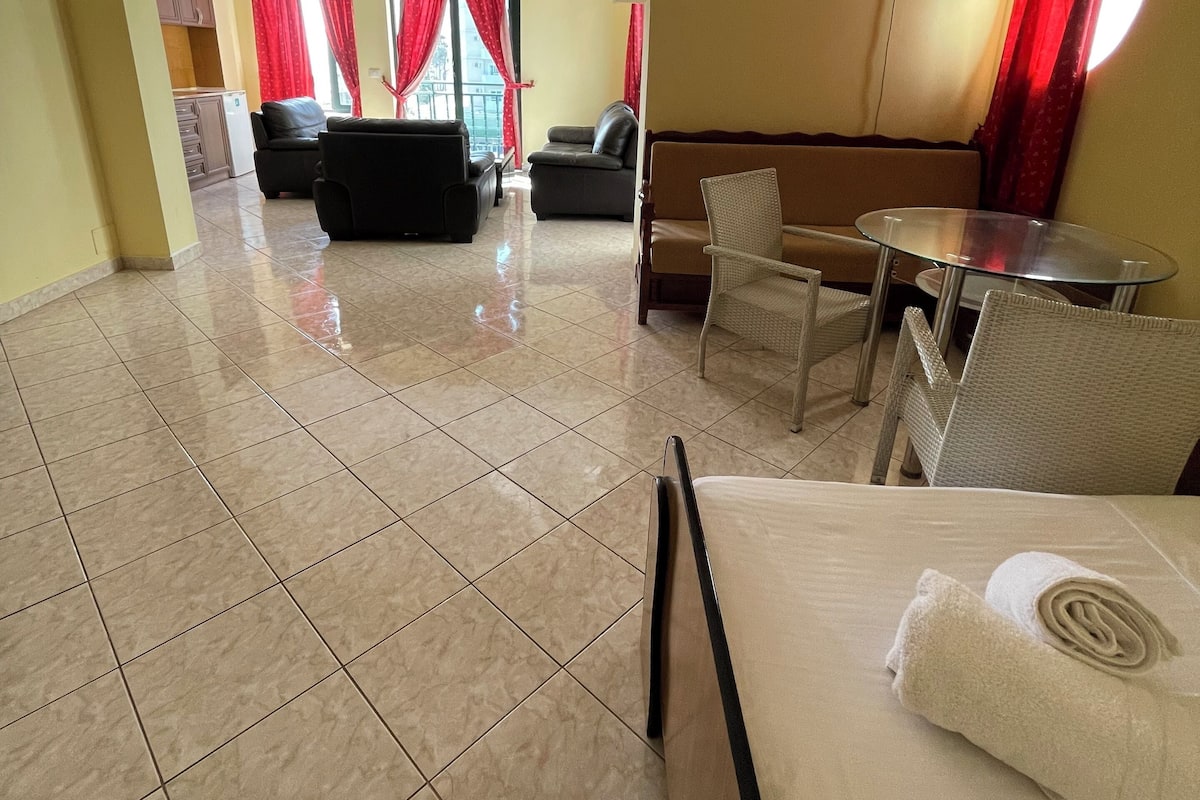 Quarto de hotel com cama, uma mesinha, um sofá e uma mesa de jantar. É visível uma área de cozinha através de um arco.