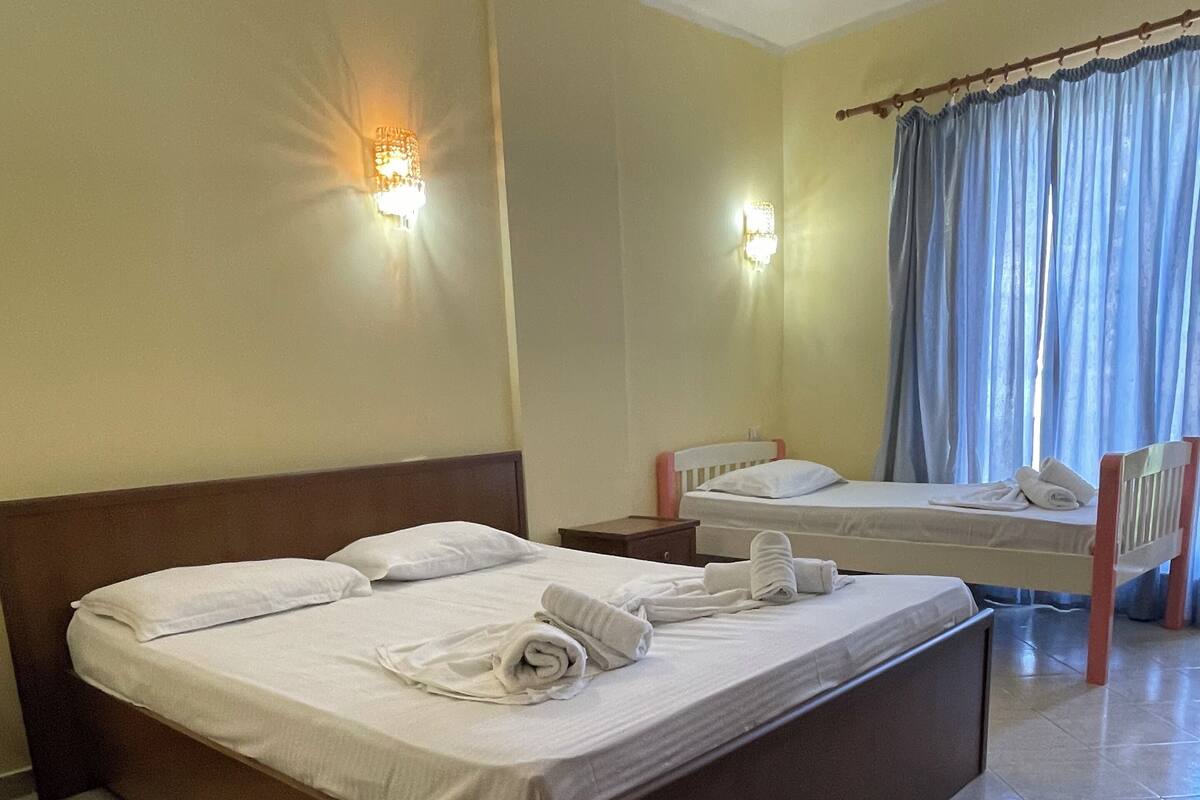 Quarto de hotel com uma cama grande, uma cama menor, uma cadeira e uma janela com cortinas.