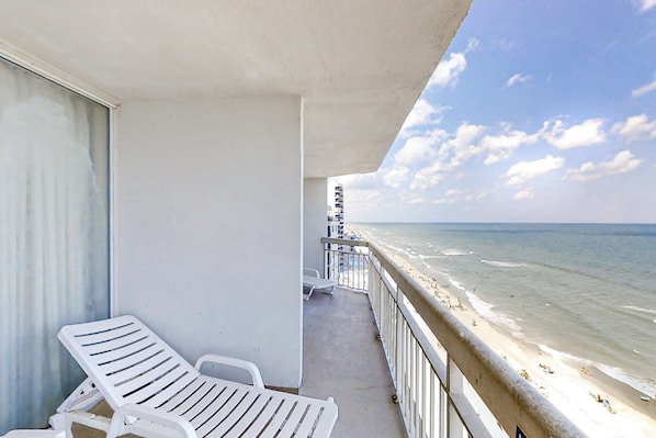 A balcony with white lounge chairs, a view of the beach, and a clear sky.