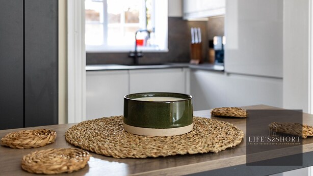 A kitchen with a green bowl on a woven placemat, a window, and a kitchen counter with bottles. - 11 Friernhay Court (Exeter)