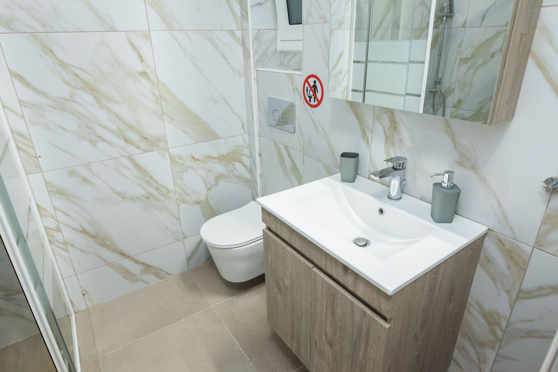 A bathroom with a marble-tiled wall, a white toilet, a sink with a wooden cabinet, and a shower area.