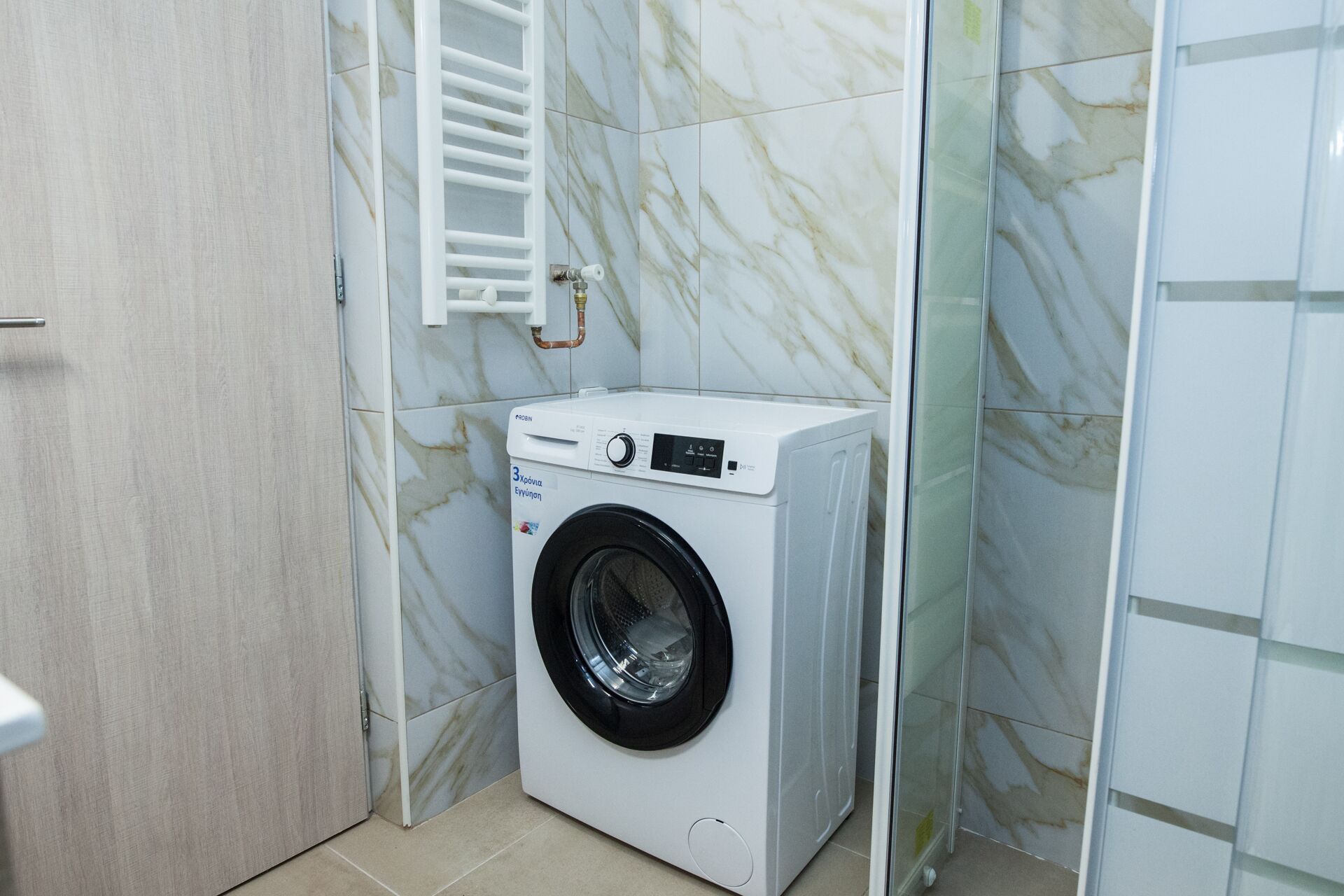 A compact, modern laundry room with a front-loading washing machine, a white towel rack, and a wooden door.
