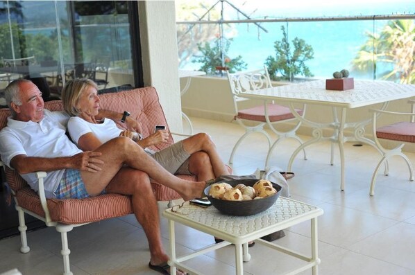 A couple sitting on a striped sofa with a table and chairs nearby, a bowl of snacks on the table, and a view of the sea through large windows.