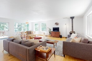 A spacious living room with a grey sofa set, a wooden coffee table, and a TV. - Water-view home with large deck, firepit, grill, & W/D (Coupeville)