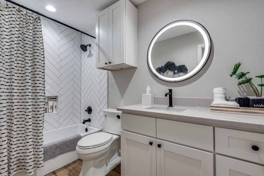 Un baño moderno con salpicadero de baldosas en espiga, una bañera independiente, un inodoro, un mueble de tocador con cajones, un espejo redondo con luz y una cortina de ducha.