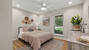 Une chambre à coucher comprenant un lit, un ventilateur de plafond, une fenêtre avec des stores, une table de chevet avec une lampe et une œuvre d’art accrochée au mur.