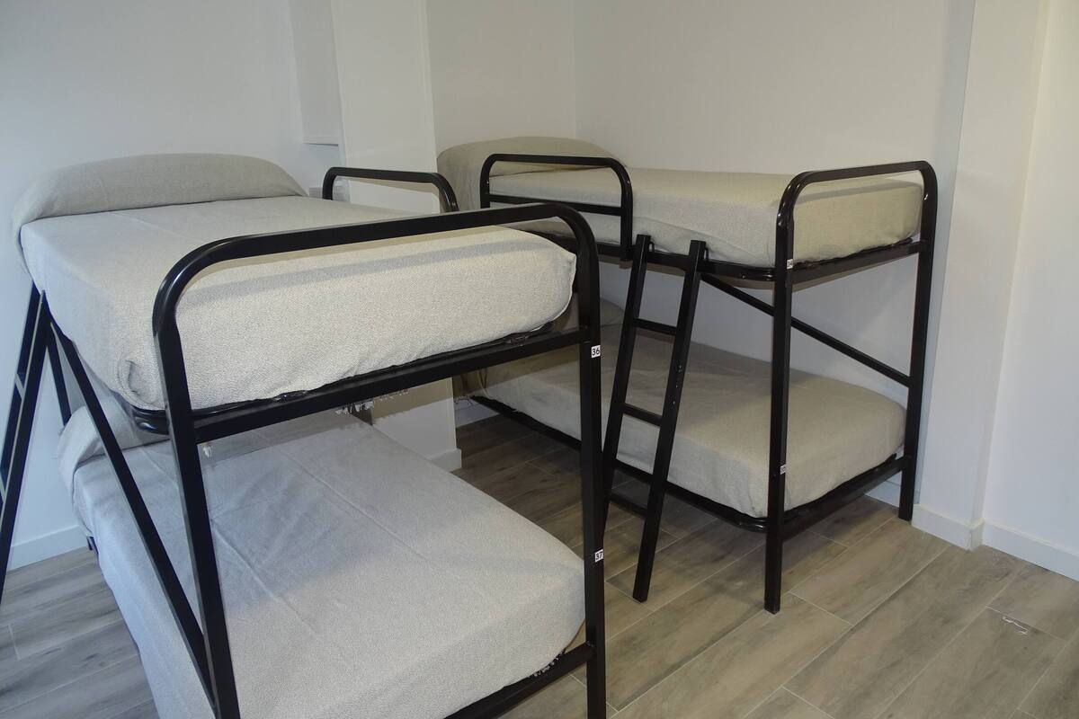 Camas literas con almohadones grises y estructuras metálicas negras en una habitación con piso de madera y paredes blancas.