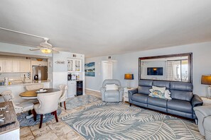 A modern living room with a blue sofa, a dining area with a round table and chairs, a kitchen with white cabinets, and a ceiling fan.