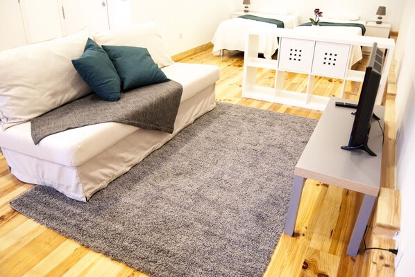 A modern hotel room with a white sofa, a grey rug, a TV on a wooden stand, and a white bedside table with a lamp.