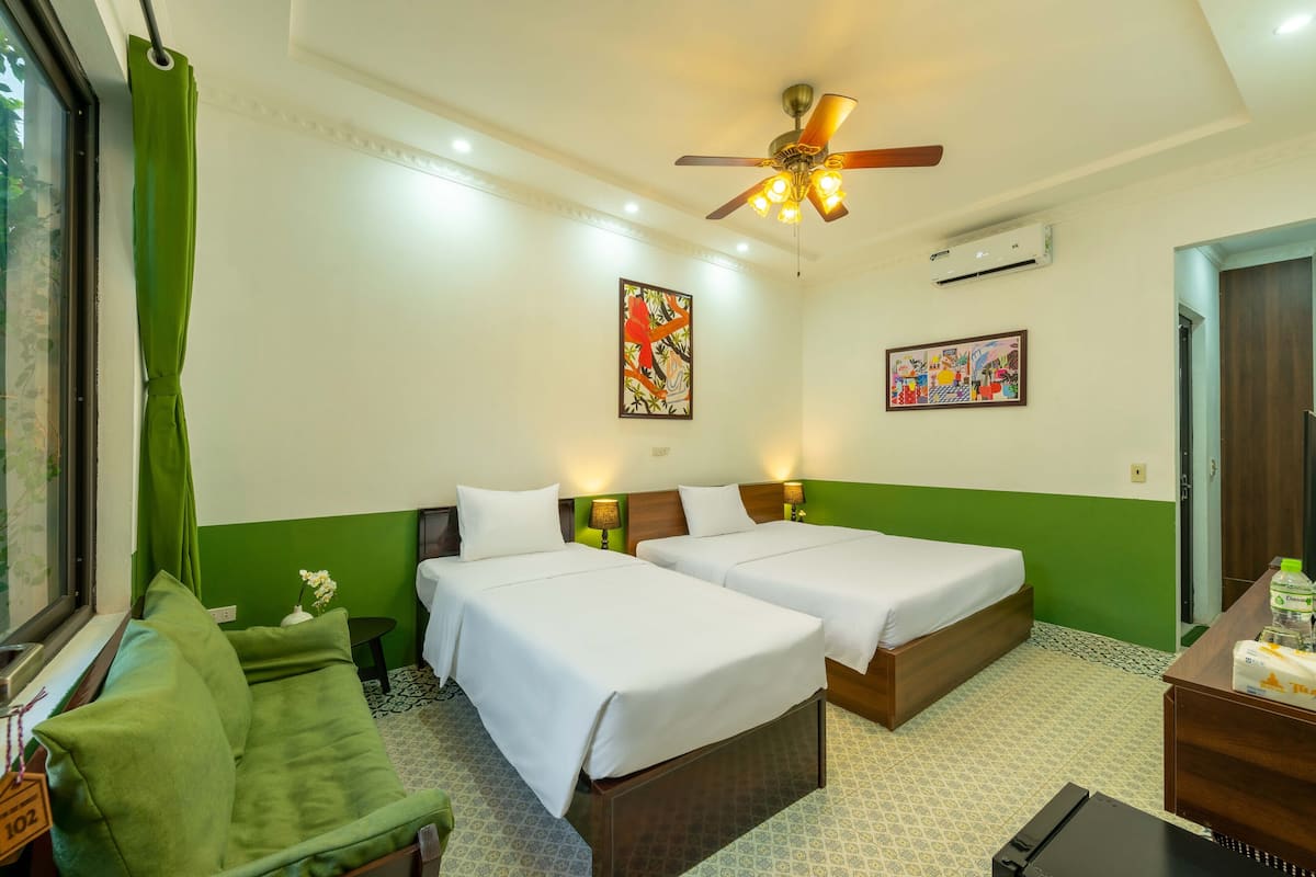 Une chambre d’hôtel avec deux lits, un canapé vert, un ventilateur de plafond et des œuvres d’art accrochées aux murs.