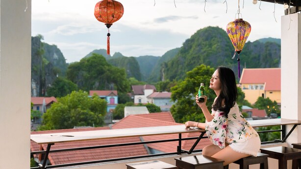 Un balcon offrant une vue sur un paysage montagneux, une personne tenant une bouteille et des lanternes traditionnelles.