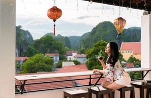 A balcony with a view of a mountainous landscape, a person holding a bottle, and traditional lanterns. - TAM COC MOMENT HOMESTAY (Nam Hoa Lu)