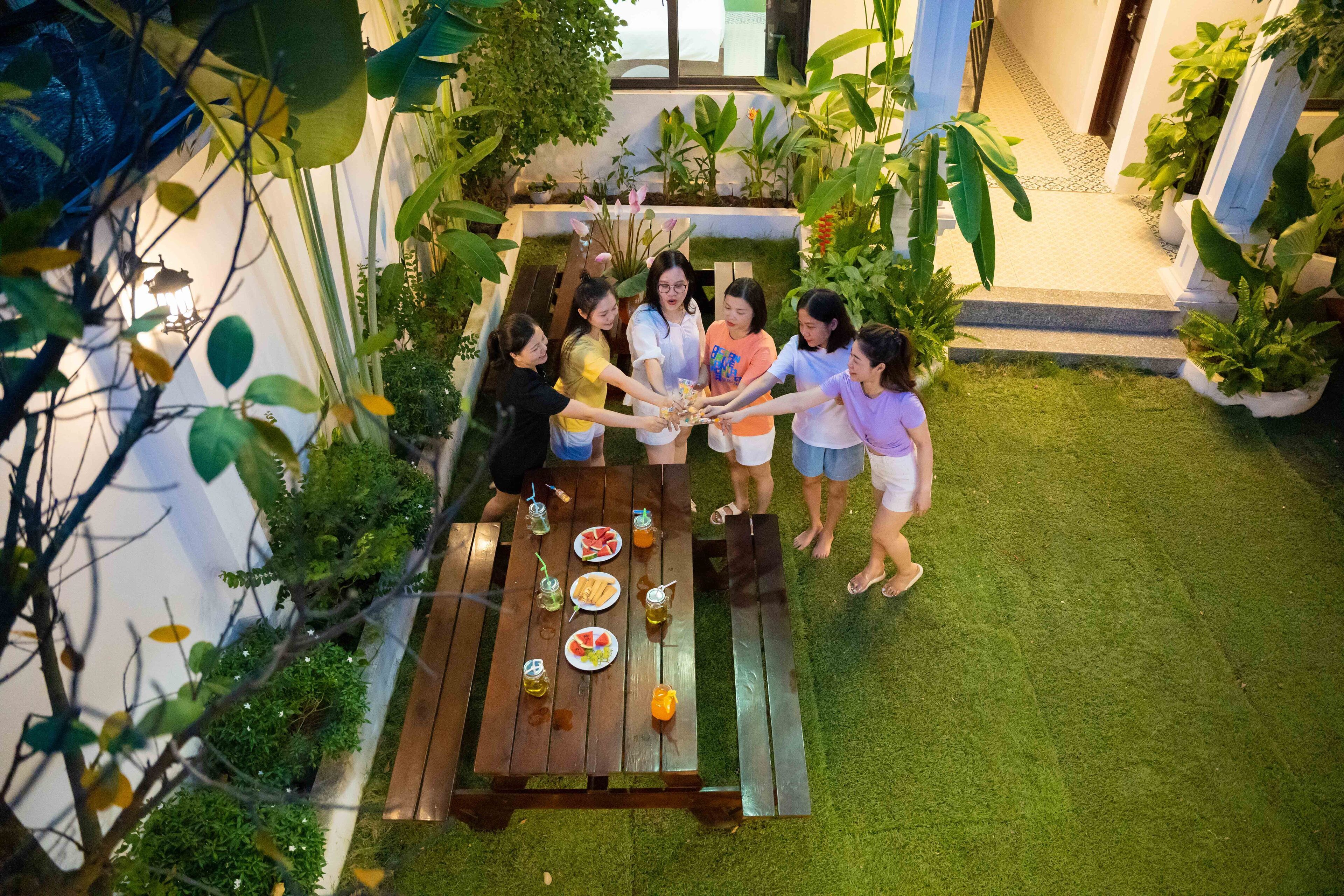 Un grupo de personas reunidas alrededor de una mesa de madera con comida y bebidas, rodeado de follaje y plantas en macetas.