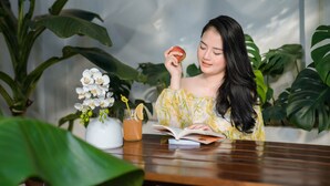 Une personne est assise à une table en bois, un livre ouvert devant elle, une plante et un verre rempli de boisson à ses côtés.