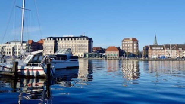 A marina with several boats docked, a prominent building with a red roof, and a clear blue sky. - Stadtnah, Eigener Garten, Ruhig (Emden)